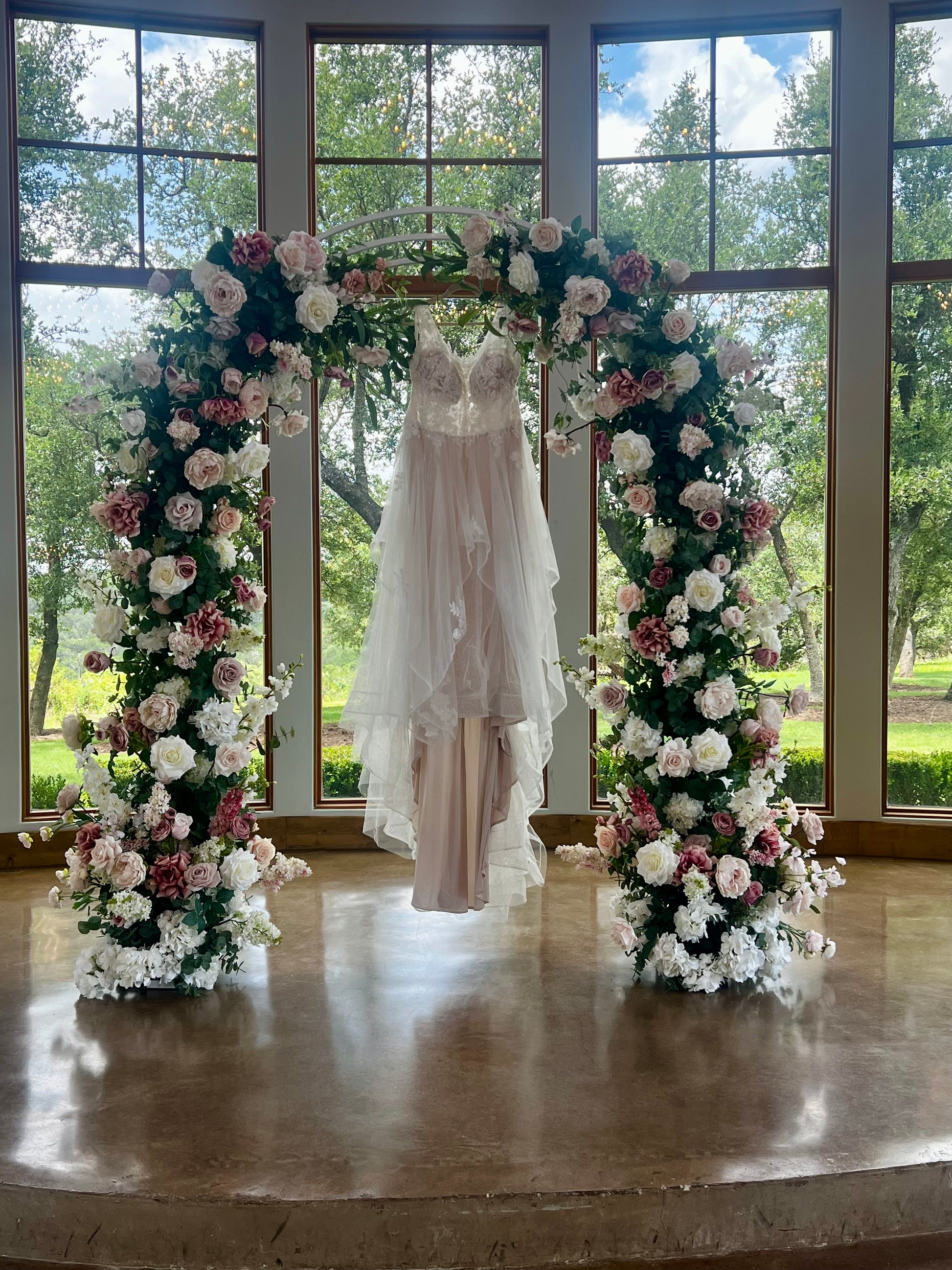 A floral arch made of faux flowers in shades of pink and blush, arranged to frame a wedding dress displayed in the center, with the arch set against a large window with a view of greenery outside.