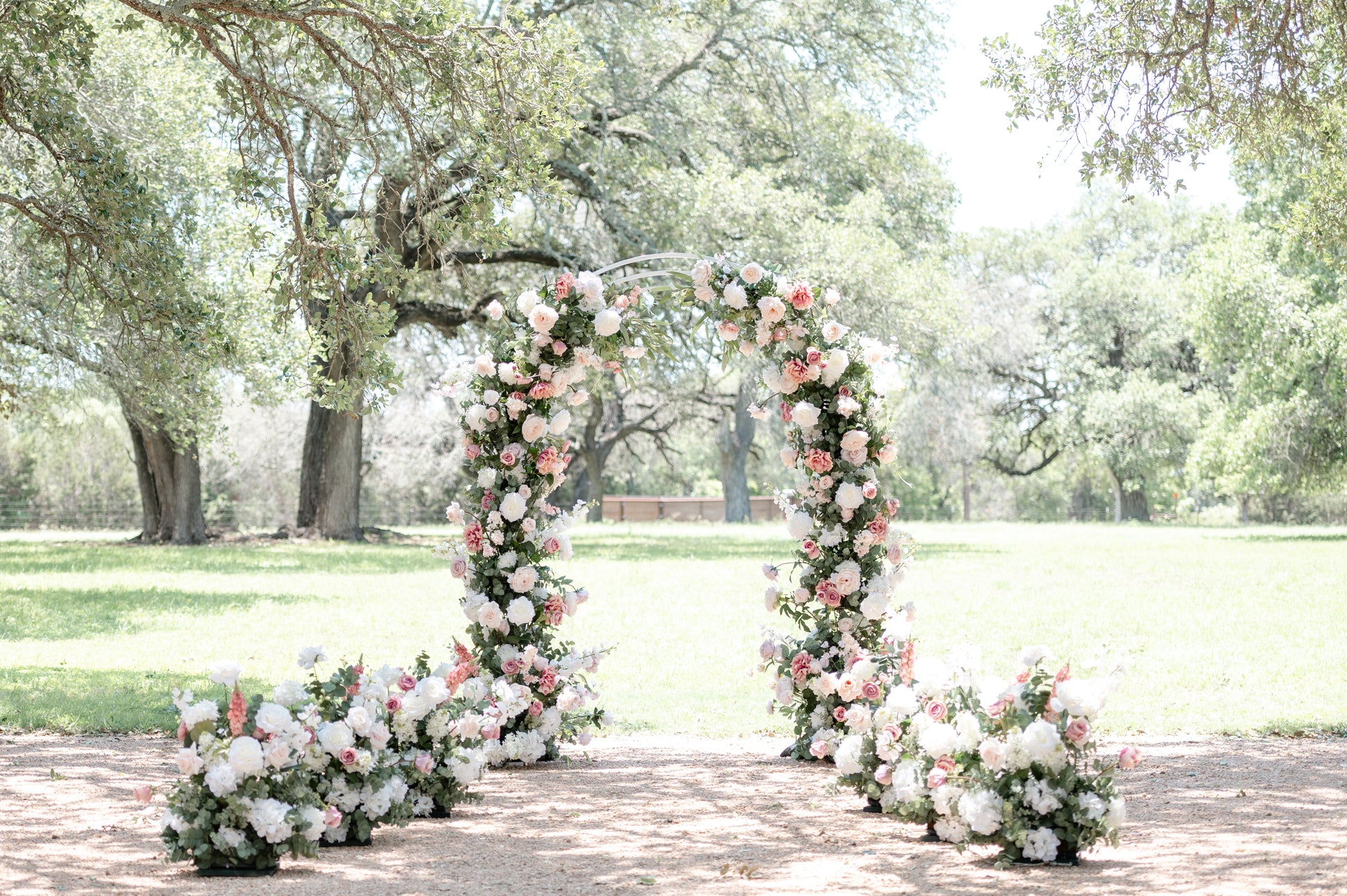 A pink and white floral arch with greenery, set in an outdoor location with trees in the background.