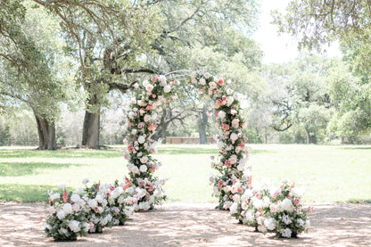 A pink and white floral arch with greenery, set in an outdoor location with trees in the background.