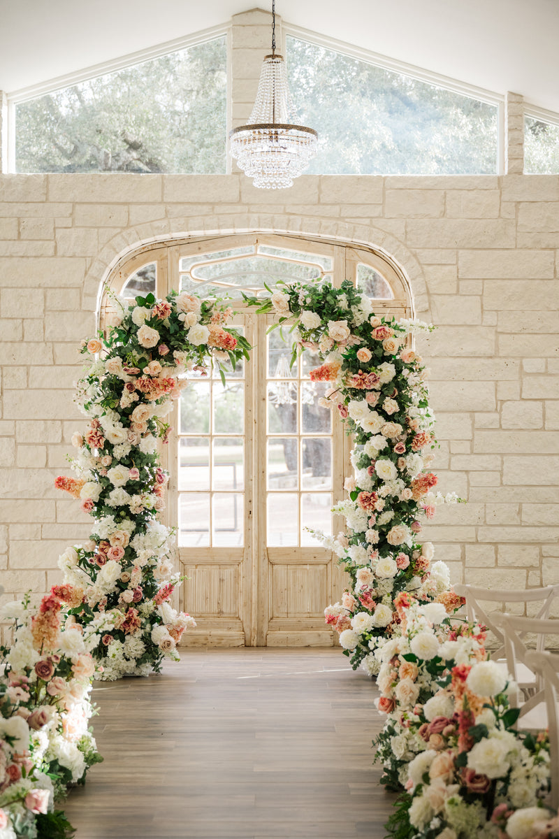 Floral archway decorated with white and pink flowers in a room with large windows and a chandelier. Venue is Lone Star Oaks in thier chapel. 
