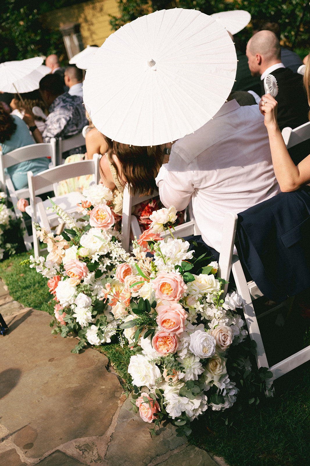 Decorative floral arrangement with white and pink flowers on a stone path, surrounded by people with parasols.