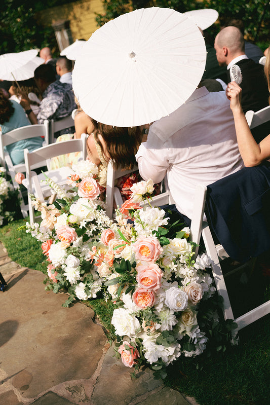 Decorative floral arrangement with white and pink flowers on a stone path, surrounded by people with parasols.