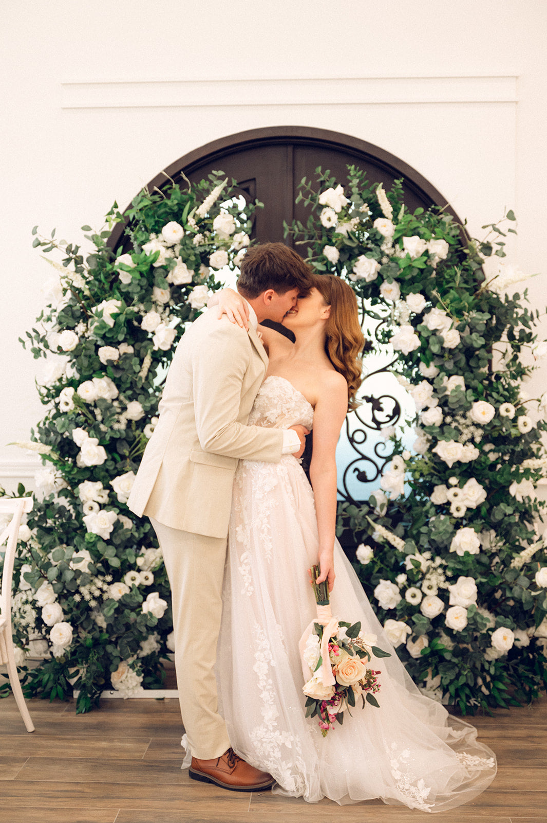 Bride and groom kissing in front of a greenery based archway